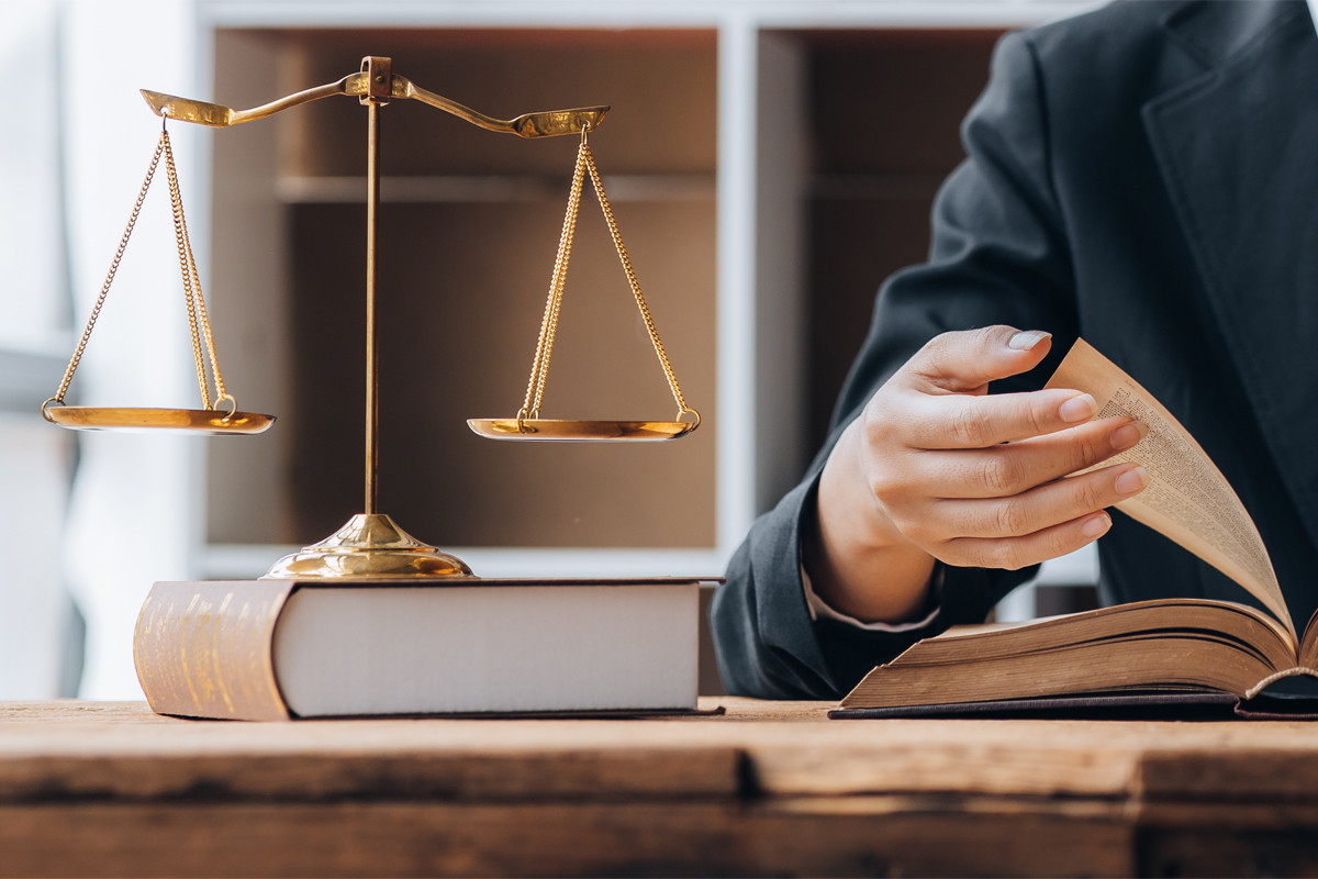 Lawyer at desk reading a law book, flipping the page, next to the scales of justice, on top of a closed law book.